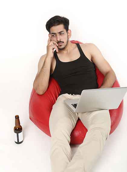 A photo of a man sitting on a bean bag with his laptop, with a bottle of beer beside him